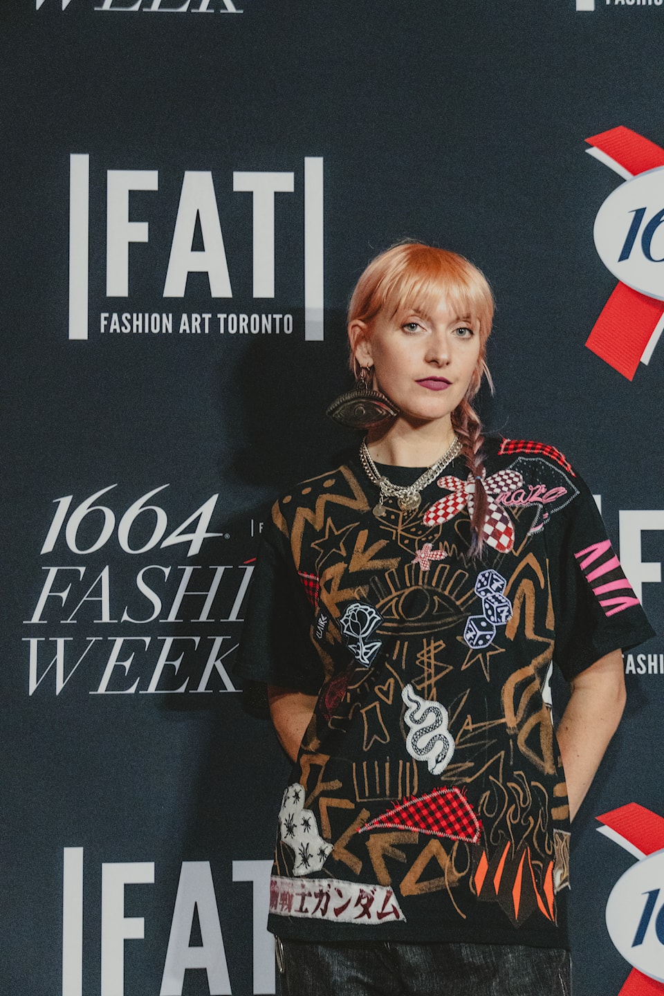 A woman waist up posing for a picture at Toronto fashion week wearing a patchwork T-shirt and a large eyeball earring.
